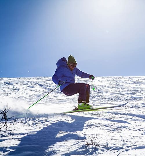 Projects A snowboarder riding down a bright, sunlit snowy slope with mountain peaks in the background."
